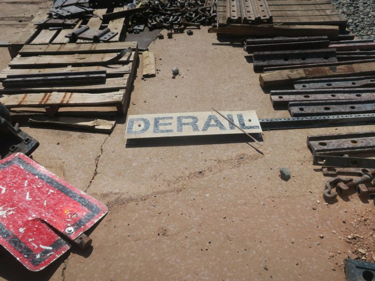 Schild mit der Aufschrift "DERAIL" auf dem Boden zwischen Schienen und Holzplatten.